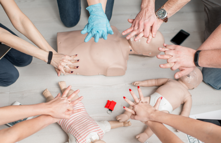 BLS and Anaphylaxis Update Hands performing CPR on training mannequins in a first aid class.